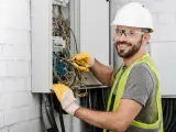Uśmiechnięty elektryk w kasku i kamizelce pracuje nad instalacją elektryczną w domu, dbając o bezpieczeństwo i porządek.