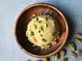 Cremoso helado de pistacho servido en un cuenco de madera, adornado con trozos de pistacho y acompañado de más pistachos enteros esparcidos alrededor.