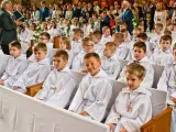 Dzieci w bieli, gotowe na rocznicę komunii świętej, siedzą w kościele. W tle widać zgromadzonych gości i fotografa.