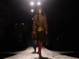 Modella su passerella, con gonna dorata e camicia marrone, un omaggio all'eleganza senza tempo di Prada, fondatore di un impero della moda.