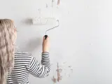 Mujer con cabello rubio y camiseta a rayas está a punto de empapelar pared con rodillo blanco.