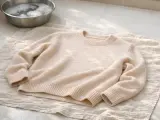 Un pull en laine beige est posé sur une serviette, prêt à laver la laine. Un bol d'eau savonneuse est à proximité.
