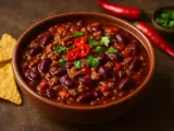 En skål med mustig chili con carne, toppad med färsk koriander och chiliskivor. Tortillachips och hela röda chilifrukter ligger bredvid.