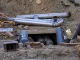 Metalowe rury tworzą skomplikowane przyłącze kanalizacyjne w wykopie. Widać fragmenty izolacji i gruzu.