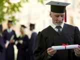 Starszy pan w todze z dyplomem zastanawia się, czy studia liczą się do stażu pracy. W tle inni absolwenci.