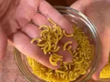 Mano sosteniendo fideos cortos y curvos sobre un bol de fideos similares.