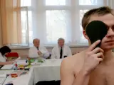 Młody mężczyzna zasłania oko podczas badania wzroku. W tle lekarze i urzędnicy pracują, ustalając, ile trwa komisja wojskowa.