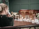 Kobieta ociera łzy chusteczką przy trumnie, otoczonej kwiatami i świecami. W tle widać wyrazy współczucia po niemiecku.