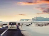 A silver van leads a convoy of vehicles driving in Iceland in December, past snow-covered mountains under a dramatic sunset sky.