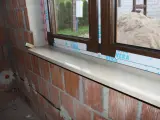 Montaż parapetów zewnętrznych po ociepleniu. Nowy, jasny parapet lśni pod brązową ramą okna, a ceglana ściana czeka na dalsze prace.