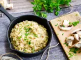 Kremowe risotto z grzybami w czarnej patelni, ozdobione natką pietruszki, obok świeże grzyby na desce.