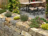 Natürliche Steinmauer mit Kräutern und Blumen, die schöne Ideen für den Garten bietet. Eine Sitzgruppe lädt zum Verweilen ein.
