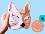 Mano sostiene un antifaz personalizado de zorro, con detalles rojos y dorados. Círculos con texto "Crea tu máscara" y un asterisco.