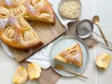 Ein köstlicher veganer Apfelkuchen, bestäubt mit Puderzucker, wird auf einem Holzbrett präsentiert. Daneben liegen frische Apfelstücke und eine Schale mit Mandelblättchen.