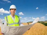 Inżynier w kasku i kamizelce odblaskowej trzyma plany na tle pola. To wizualizacja, jak działka rolno budowlana co to znaczy, czyli połączenie upraw z możliwością zabudowy.
