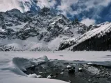 Zamarznięte Morskie Oko, otoczone ośnieżonymi szczytami Tatr. W oddali widać ludzi spacerujących po lodzie, a na pierwszym planie płynie strumyk.