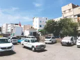 Taxis warten auf Fahrgäste in einer sonnigen Stadt in Marokko. Weiße Gebäude und Bäume säumen die Straße.
