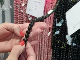 Manos con uñas rojas sostienen un collar de cuentas negras de shungita. En el fondo, se ven hilos de cuentas rosas y blancas.