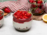 Zwei Gläser mit cremigem Erdbeer-Cheesecake, garniert mit frischen Erdbeeren und Erdbeersauce. Daneben ein Glas voller Erdbeeren und eine Zitronenhälfte.