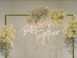 Elegancka ściana z kwiatów w kremowych odcieniach, ozdobiona neonowym napisem "Better Together".
