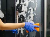 Main bleue, éponge orange pour nettoyer paroi douche. Mousse et bulles sur la paroi vitrée.