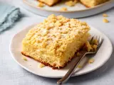 Ein Stück saftiger Streuselkuchen vom Blech, bestäubt mit Puderzucker, liegt auf einem Teller. Eine Gabel wartet darauf.