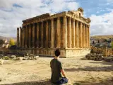 A man sits contemplating the grandeur of Roman ruins in Lebanon, bathed in sunlight under a cloudy sky.