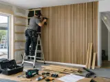 Man på en stege, som ska montera akustikpanel på väggen med en omladdningspistol. Verktyg och material ligger utspridda på golvet.