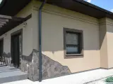 Elewacja domu w kolorze kawa z mlekiem, ozdobiona kamiennym cokołem. Widoczne okna i drzwi tarasowe.