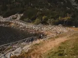 Dwie osoby idą kamiennym szlakiem przy jeziorze, otoczonym drewnianym ogrodzeniem. W oddali widać las i góry.