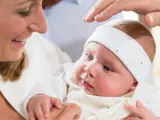 Uśmiechnięta mama trzyma dziecko w białej szatce. Dłoń ojca błogosławi malucha. Zastanawiasz się, ile kosztuje chrzest?