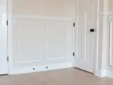A room with a DIY chair rail, wainscoting, and white doors. The walls are painted a light cream color.