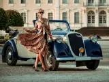 Elegancka kobieta w kwiecistej sukience i okularach przeciwsłonecznych opiera się o zabytkowy samochód. Stylizacja nawiązuje do mody lat 20.