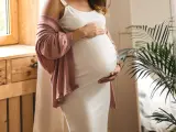 Frau in weißem Kleid und rosafarbenem Cardigan hält ihren Bauch, 34 ssw. Ein Fenster und eine Pflanze sind im Hintergrund.