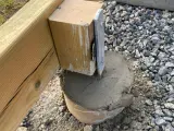 En trästolpe fästs i en betongplint, en del av arbetet med att bygga altan på plintar. Grus och jord syns runt plinten.