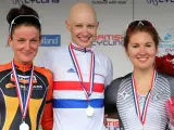 Joanna Rowsell, a bald cyclist in a white and red jersey, smiles on the podium with two other medalists.