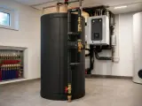 Duży, czarny bufor ciepła w kotłowni, obok białego zasobnika i kotła. Widać instalację hydrauliczną.