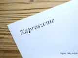Eleganckie zaproszenie na ślub na białym, satynowym papierze, leżące na drewnianym tle.