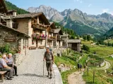 Un escursionista cammina in un pittoresco borgo del Trentino Alto Adige, con case in pietra e fiori colorati, mentre sullo sfondo si stagliano maestose montagne.