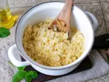 Ein Holzlöffel lockert den frisch zubereiteten Couscous in einem Topf auf. Daneben stehen Öl und Minze.