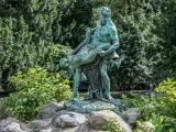 Bronzestatue eines Mannes, der eine Frau trägt, vor einem grünen Hintergrund mit einem kleinen Wasserfall in Berlin.