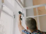 Mężczyzna przykręca zawias do okna. Planujesz wymianę okien w bloku? Cena zależy od wielu czynników.