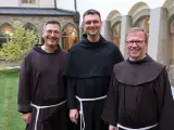 Drei Franziskaner Minoriten in braunen und schwarzen Kutten lächeln in die Kamera vor einer Klosterfassade.