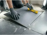 Ręce w rękawiczkach układają szarą płytkę, sprawdzając poziom. To kluczowy moment, jak zacząć układanie płytek na podłodze, by efekt był idealny.