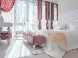 Elegancka sypialnia w stylu angielskim z pikowanym zagłówkiem, różowymi pufami i zasłonami.