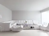 Soggiorno minimalista con divano componibile bianco, tappeto grigio e tavolino trasparente. Le tonalità di bianco per pareti creano un'atmosfera eterea.