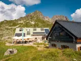 Die Buchel Alpe, eine Berghütte mit Solaranlagen, liegt vor einer beeindruckenden Bergkulisse. Gäste sitzen draußen unter Sonnenschirmen.