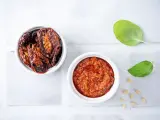 Due ciotoline bianche: una con pomodori secchi interi, l'altra con un invitante pesto di pomodori secchi. Foglie di basilico e pinoli completano la scena.