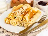 Ein Teller mit goldbraunen Quark Pfannkuchen, aufgerollt und gestapelt. Daneben liegen ein goldenes Messer und eine Gabel. Im Hintergrund eine Schale Marmelade.