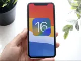 Dłoń trzyma iPhone'a z ikoną iOS 16 na kolorowej tapetcie.